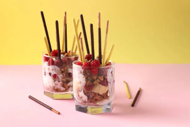 Photo of Dessert in two glasses with chocolate granola, banana and raspberry, decorated with sticks Pocky on yellow and pink background