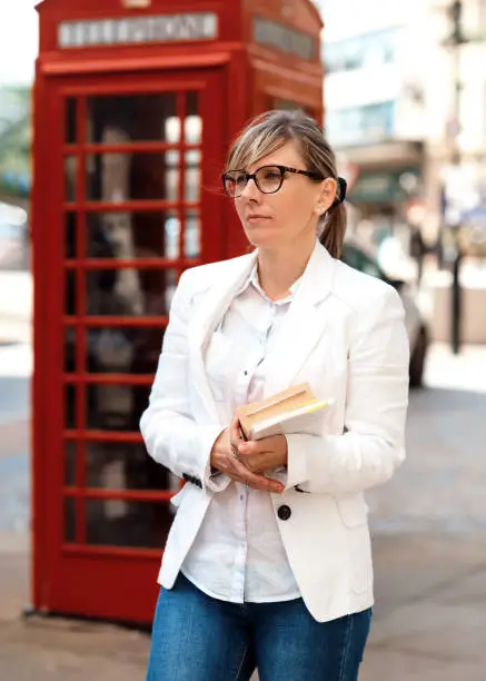 A woman wearing glasses, a white coat, jeans and white shirt holding books against the red telephone box in CityCenter