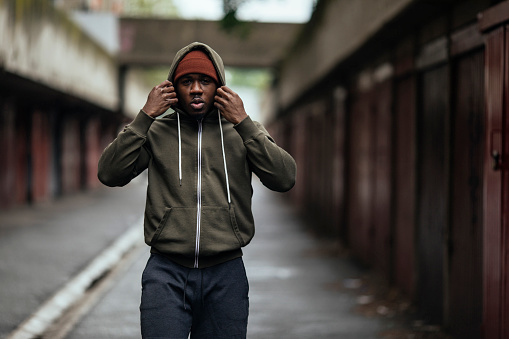 Young man with a hat on putting on hood after finishing a workout in a street alley