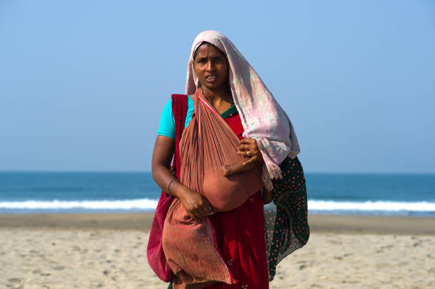 Mother with a baby in her arms, covered from the sun with a cloth, begs for alms on the beach in Goa Goa, India, February 14, 2011. A young woman asks for money to support her baby on a beach in the Indian state of Goa. in India, a significant number of people live below the poverty line and do not even have a roof over their heads. Therefore, asking for money on the street for many is the only way to survive. walking beach women sadness stock pictures, royalty-free photos & images