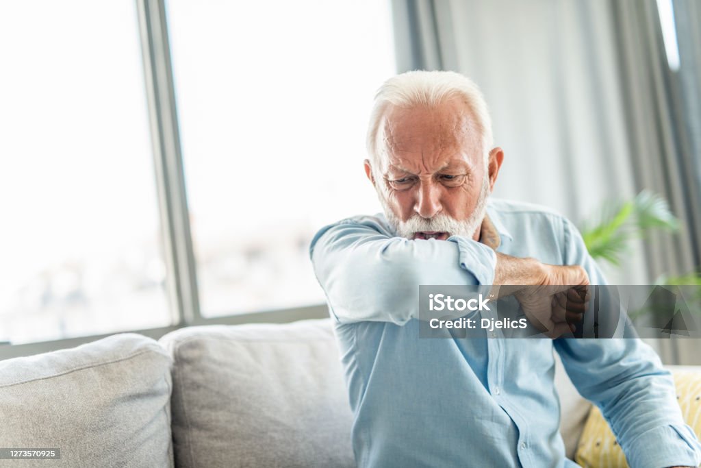 Senior man coughing. Front view of senior man coughing into elbow at home. Coughing Stock Photo