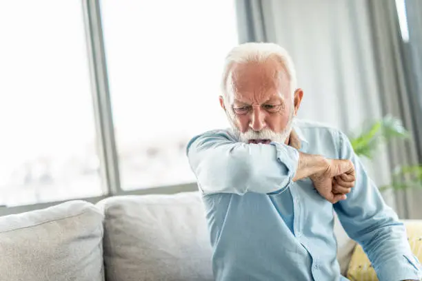 Front view of senior man coughing into elbow at home.