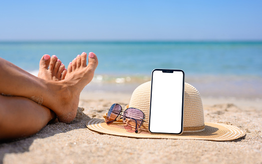 Smartphone with blank white display on the beach. Hat, sunglasses and legs of the girl on the background of the sea. The concept of using gadgets and the Internet at a seaside resort.