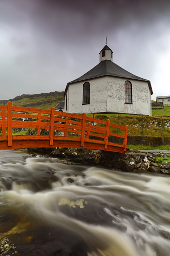 Fisheye lowangle shot of Haldorsvíkar Church with orange bridge and small river taken with long exposure on a cloudy rainy day in Haldorsvik, Faroe Islands.