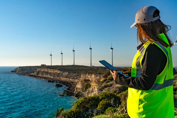 화창한 날에 풍력 터빈에 대한 디지털 태블릿을 들고 서있는 여성 엔지니어가 풍력 터빈을 보고 있습니다. - energy fuel and power generation wind turbine outdoors 뉴스 사진 이미지