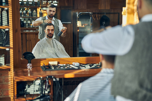 Enthusiastic young barber spraying hair of his client with a fixator while giving him a new look