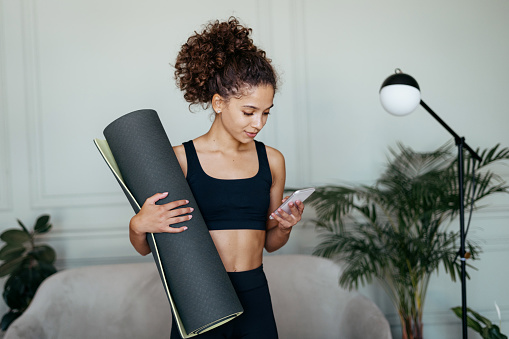 Smiling adult sporty afro american woman using online app on modern smartphone, holding yoga mat in hand, prepare training at home, making morning sport exercise