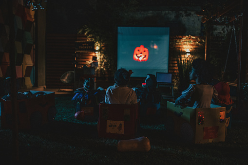 Group of children at backyard movie night. Sitting in cardboard box cars, eating popcorn and watching movie. Social distance during Covid-19 pandemic