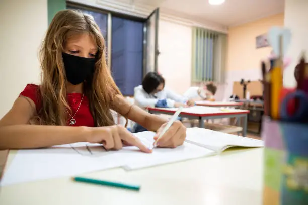 Young Caucasian schoolgirl wearing a protective face mask in the classroom while working on schoolwork at her desk.