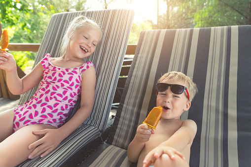 Two cute adorable caucasian blond little siblings enjoy having fun relaxing and eat fruit popsicle icecream sitting on sunbed in yard garden near pool sea beach. Brother and sister chilling outdoor.