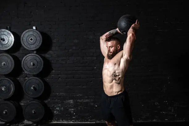 Young sweaty strong muscular fit man with big muscles doing ball throwing on the floor as hardcore cross workout training in the gym