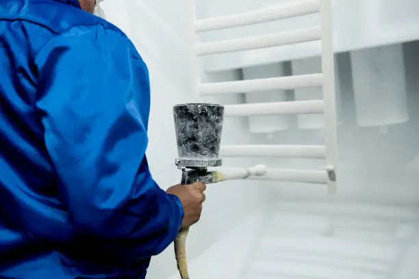 Worker wearing protective wear performing powder coating of metal details in a special industrial camera