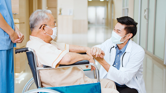 Young male Asian doctor crouch down holding hand and talk to the senior adult patient on wheelchair in hospital hallway. Medical healthcare job, or hospital business concept