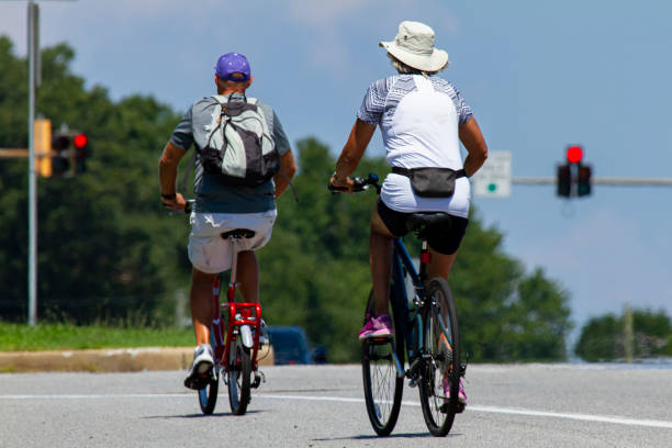 晴れた夏の日には、カジュアルなショートパンツやtシャツ、帽子を着た先輩カップルが高速道路の近くで自転車に乗っています。 - senior adult action cycling senior couple ストックフォトと画像
