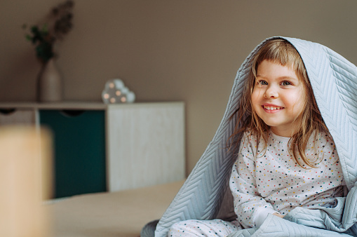 Happy beautiful little girl having fun time lying on the bed under the blanket in the bedroom.