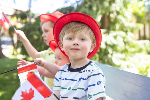 Three young siblings sit on a park bench and wave Canadian flags Two cute young brothers relax on a bench, with their little toddler sister between them, and wave small Canadian flags in the air. They're all wearing hipster brimmed hats and having a wonderful time on a summer afternoon. 6-11-months stock pictures, royalty-free photos & images