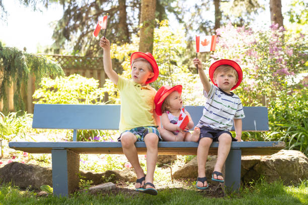 Three young siblings sit on a park bench and wave Canadian flags Two cute young brothers relax on a bench, with their little toddler sister between them, and wave small Canadian flags in the air. They're all wearing hipster brimmed hats and having a wonderful time on a summer afternoon. 6 11 months stock pictures, royalty-free photos & images