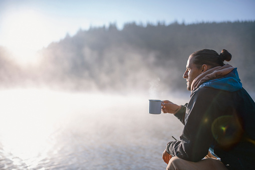Side view of kneeling man drinking coffee or tea on wooden pier around beautiful foggy mountain lake at sunrise.