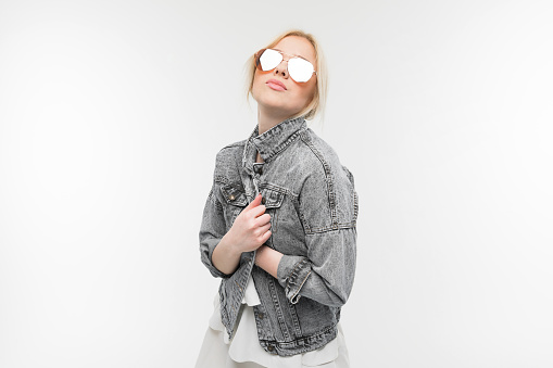 portrait of young charming joyful blonde girl in jeans jacket possing and gesticulating in round sunglasses on background