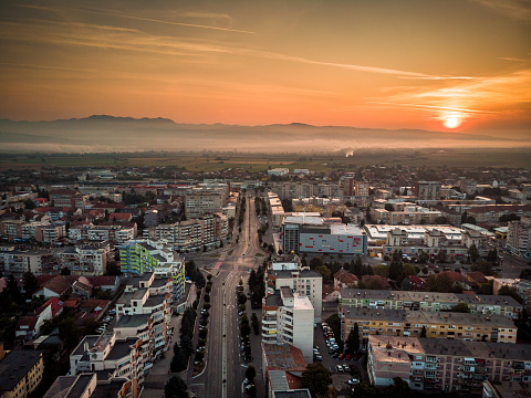 Color image depicting a high angle drone view of the city of Deva, a town in the Transylvania region of Romania. We can see communist-style apartment blocks and Deva's ancient citadel in the distance. The scene is set off by a sunset and cloudscape.