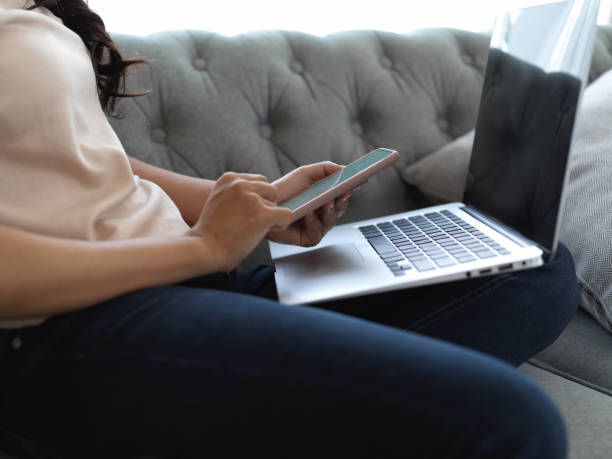 istock Female using smartphone while working with laptop in living room 1273797888