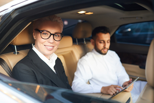 Mature businesswoman in eyeglasses smiling at camera while sitting in the car with his colleague in the background