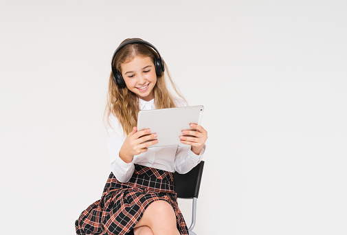 Cute smiling teen girl in school uniform with headphones and tablet in her hands isolated on white background