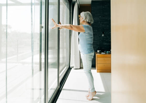 Elderly woman standing on tiptoe in bedroom Side view of elderly female exercising at home. Full length of senior woman with white hair is looking through window. She is standing on tiptoe in bedroom. tiptoe stock pictures, royalty-free photos & images