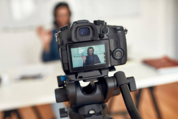 istock Hello. Young happy male teacher looking at camera and waving while giving online class through webcam at home. Tutor recording video tutorial. Focus on a camera screen 1273817993