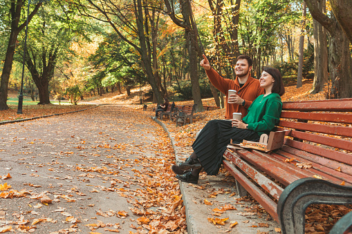 couple sitting at public park bench at autumn sunny day pointing at copy space. drink to go