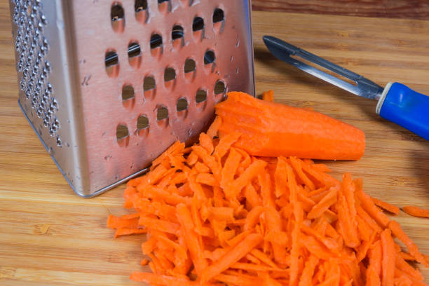 Pile of grated carrot in large slices against the stainless steel kitchen grater on the wooden cutting board, close-up in selective focus