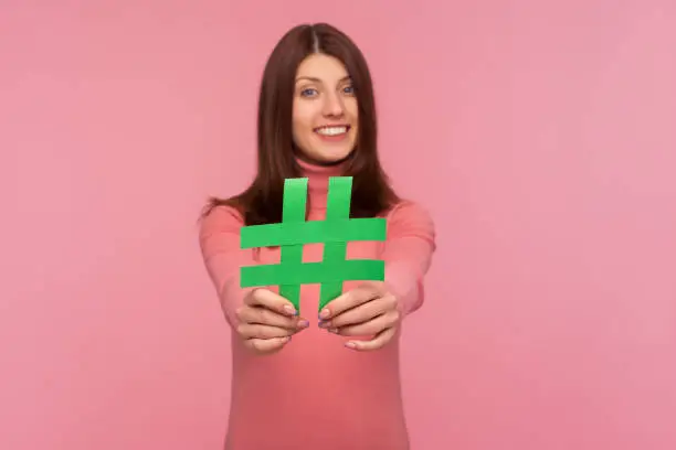 Photo of Cheerful positive female blogger in pink sweater holding green hashtag sign in hands, easy blog search, tagging