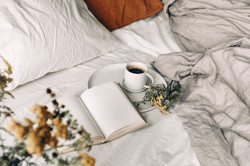 Fall breakfast still life. Cup of coffee, open book and tansy flowers on white linen background. Blurred floral foreground Velvet and linen pillows in bed. Moody autumn lifestyle concept. Top view.