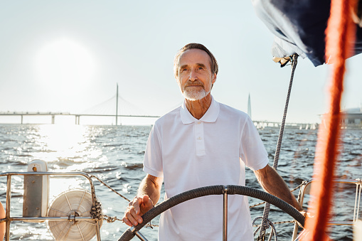 Mature man steering a private yacht while on vacation. Bearded yachtsman standing on his sailing boat looking at distance.