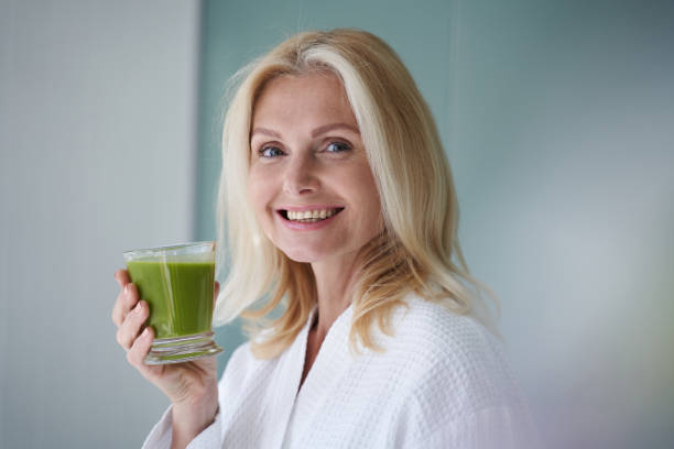 Happy blonde lady standing with healthy drink Waist up of smiling attractive adult woman going to drink green smoothies while holding glass against wall 40 49 years stock pictures, royalty-free photos & images