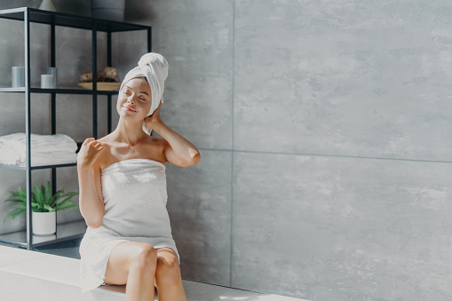 Relajado joven modelo femenino caucásico lleva toalla envuelta en la cabeza, se siente refrescado después de tomar la ducha, tiene piel suave y limpia y saludable, posa en el baño acogedor. Concepto de mujeres, belleza e higiene. photo
