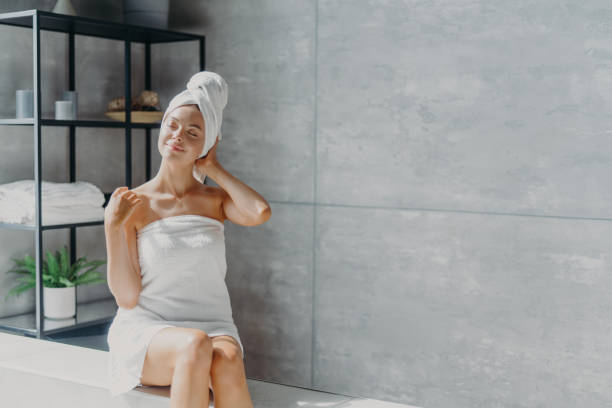 Relaxed young Caucasian female model wears towel wrapped on head, feels refreshed after taking shower, has healthy clean soft skin, poses in cozy bathroom. Women, beauty and hygiene concept. Relaxed young Caucasian female model wears towel wrapped on head, feels refreshed after taking shower, has healthy clean soft skin, poses in cozy bathroom. Women, beauty and hygiene concept. woman wrapped in bath towel stock pictures, royalty-free photos & images