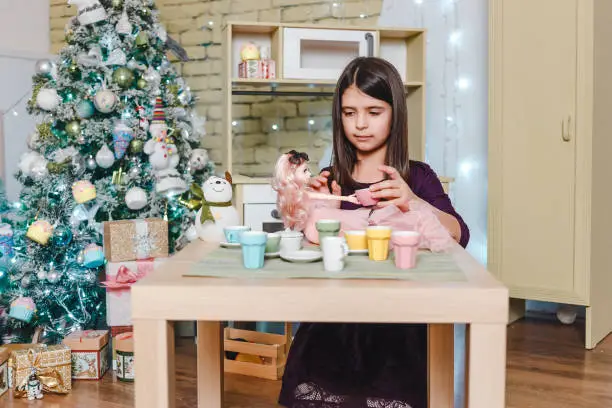 Seven-year-old beautiful dark-haired girl plays with doll at tea party table in Christmas room
