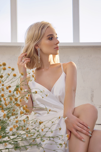 Sexy blonde woman in a beautiful white dress is sitting near the window in front of a bouquet of wild flowers. Romantic girl with beautiful natural makeup