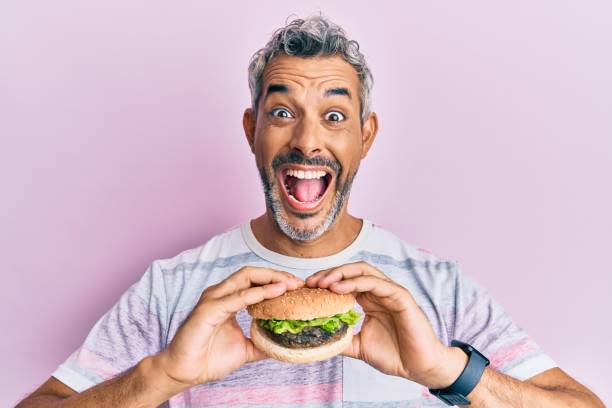 Middle age grey-haired man eating a tasty classic burger celebrating crazy and amazed for success with open eyes screaming excited. Middle age grey-haired man eating a tasty classic burger celebrating crazy and amazed for success with open eyes screaming excited. spanish hamburger stock pictures, royalty-free photos & images