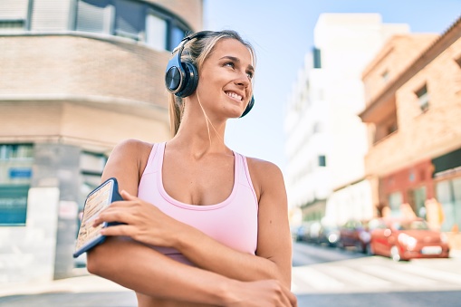 Young blonde sporty girl using headphones walking at street of city.