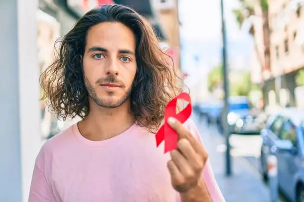 Photo of Young hispanic man with serious expression holding hiv awareness red ribbon at city.