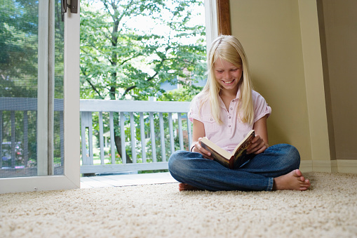Girl (7-9) sitting cross-legged reading book smiling