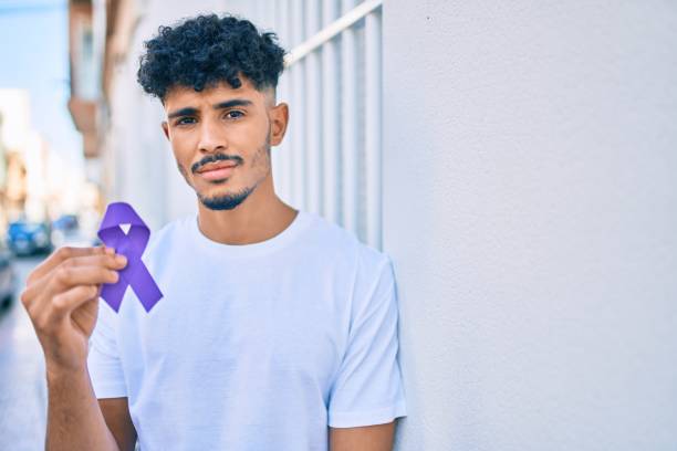 молодой арабский человек с серьезным выражением, держащим фиолетовую ленту осознания, опираясь на стену. - purple ribbon alzheimers disease alertness стоковые фото и изображения