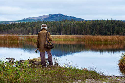 A grey haired senior man standing beside a Scottish loch. The man is wearing an old waxed jacket and a tweed cap. He is holding a fly fishing rod and has a bag over his shoulder. The loch is in a remote rural location in Dumfries and Galloway, south west Scotland. It is late summer and the plants are starting to change colour on this overcast morning.