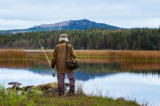 A grey haired senior man standing beside a Scottish loch. The man is wearing an old waxed jacket and a tweed cap. He is holding a fly fishing rod and has a bag over his shoulder. The loch is in a remote rural location in Dumfries and Galloway, south west Scotland. It is late summer and the plants are starting to change colour on this overcast morning.