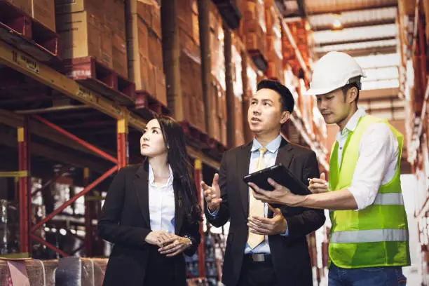 Photo of Asian male foreman or manager showing report from digital tablet to business man and woman or CEO in warehouse company. people discuss of logistics shipping, quality control or export import goods.