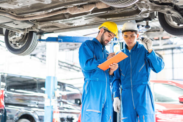 istock Caucasian automotive mechanic man writing on maintenance document clipboard and checking under car in auto repair garage, assistant pointing flash light, transport business and after service concept 1273876642