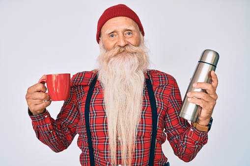Old senior man with grey hair and long beard holding cup of coffee and thermo smiling with a happy and cool smile on face. showing teeth.