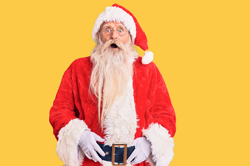 Old senior man with grey hair and long beard wearing traditional santa claus costume afraid and shocked with surprise and amazed expression, fear and excited face.